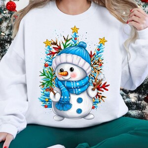 May include: White sweatshirt featuring a cheerful snowman design. The snowman wears a blue hat and scarf, surrounded by festive greenery and stars. A perfect holiday or winter season apparel.