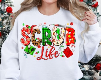 May include: White sweatshirt featuring the text "SCRUB LIFE" in a festive design. The letters are decorated with patterns like leopard print, plaid, and snowflakes, with medical-themed illustrations. Christmas trees and ornaments are in the background.