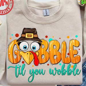 May include: A beige sweatshirt with a turkey graphic that says "GOBBLE til you wobble". The turkey has a brown hat, blue eyes, and a red wattle. Colorful polka dots surround the text. The sweatshirt has a round neck and long sleeves.