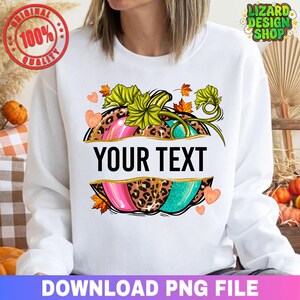 May include: White sweatshirt featuring a pumpkin design with the text "YOUR TEXT". The pumpkin is split into sections of pink, teal, and leopard print, with green leaves and orange accents. The design includes hearts and fall leaves.