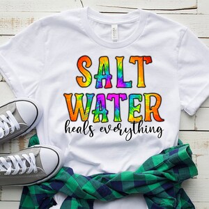 Puede incluir: Camiseta blanca con un dise&ntilde;o tie-dye que dice "Salt Water heals everything".