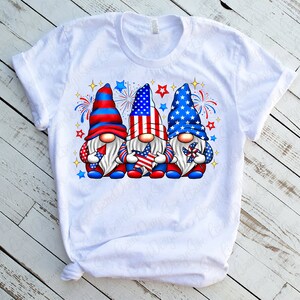K&ouml;nnte beinhalten: Wei&szlig;es T-Shirt mit einem Design aus drei Gnomen, die rote, wei&szlig;e und blaue H&uuml;te tragen und Sterne und Windm&uuml;hlen halten. Die Gnome sind von roten, wei&szlig;en und blauen Feuerwerksk&ouml;rpern umgeben.