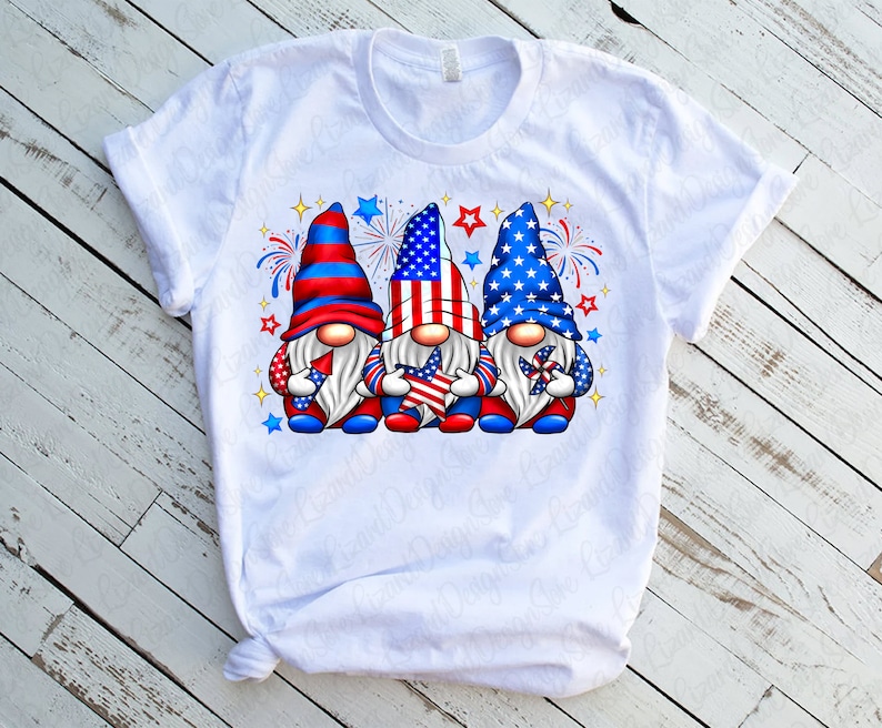 K&ouml;nnte beinhalten: Wei&szlig;es T-Shirt mit einem Design aus drei Gnomen, die rote, wei&szlig;e und blaue H&uuml;te tragen und Sterne und Windm&uuml;hlen halten. Die Gnome sind von roten, wei&szlig;en und blauen Feuerwerksk&ouml;rpern umgeben.