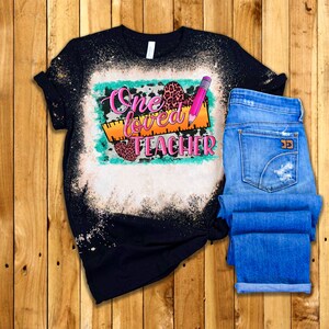 Puede incluir: Una camiseta negra con un dise&ntilde;o salpicado de lej&iacute;a que presenta el texto "One Loved Teacher" en rosa y naranja con una regla, un l&aacute;piz y corazones de estampado de leopardo.