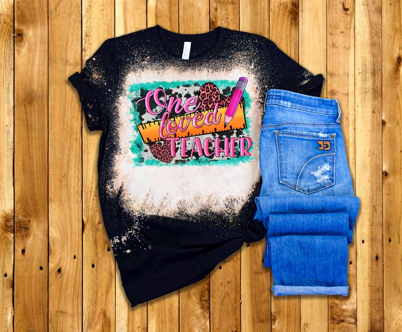 Puede incluir: Una camiseta negra con un dise&ntilde;o salpicado de lej&iacute;a que presenta el texto "One Loved Teacher" en rosa y naranja con una regla, un l&aacute;piz y corazones de estampado de leopardo.