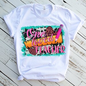 Puede incluir: Camiseta blanca con un dise&ntilde;o gr&aacute;fico que dice "One loved teacher" con un l&aacute;piz rosa y una regla, estampado de leopardo y estampado de vaca.