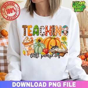 May include: White sweatshirt with a colorful graphic design featuring the word "TEACHING" in various patterns. The design includes pumpkins, sunflowers, a coffee cup, and the phrase "little pumpkins".