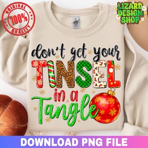 Puede incluir: Sudadera beige con la frase "don't get your Tinsel in a Tangle". La palabra "TINSEL" presenta varios diseños, incluyendo estampado de leopardo y rayas de bastón de caramelo. A la derecha, un adorno rojo.