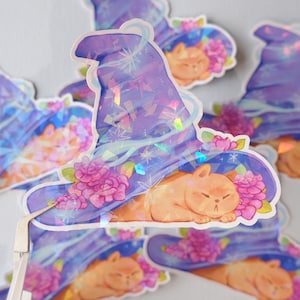 Puede incluir: Una pegatina holográfica con un gato durmiendo dentro de un sombrero de mago morado y azul con flores rosas. El sombrero tiene un acabado brillante e iridiscente.