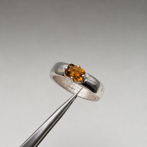 May include: A silver ring featuring a rectangular, faceted amber-coloured gemstone set in a four-prong setting. The band has a smooth, polished finish and a rounded profile, showcasing a clear contrast between the metallic silver and the warm stone.