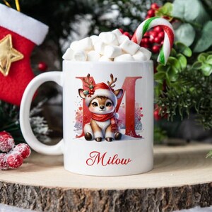 Peut inclure: Mug en céramique blanche sur le thème de Noël. La tasse présente un cerf de dessin animé portant un bonnet et une écharpe de Père Noël, la lettre "M" et le nom "Milow". La tasse est remplie de guimauves et d'un sucre d'orge.
