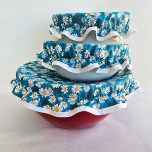 Peut inclure: Trois couvercles de bol réutilisables avec un motif de marguerite blanche sur fond bleu sarcelle. Les couvercles sont en tissu et ont un bord blanc à volants.