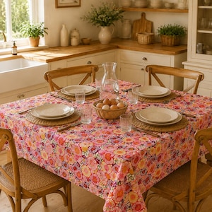 Könnte beinhalten: Ein quadratischer Esstisch mit einer Tischdecke mit pinkfarbenem, orangefarbenem und violettem Blumenmuster. Der Tisch ist mit vier runden geflochtenen Platzsets, weißen Keramiktellern, klaren Trinkgläsern, Besteck und einem Korb mit Eiern gedeckt.