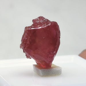 Könnte beinhalten: Ein tief roter, facettierter Edelstein mit einer rauen, unregelmäßigen Form. Der Edelstein wird auf einem weißen Schaumstoffpolster präsentiert.