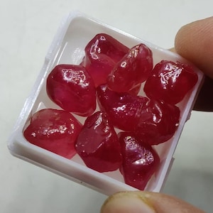 Könnte beinhalten: Eine kleine weiße Schachtel gefüllt mit losen, facettierten Rubinen. Die Rubine sind tiefrot und haben eine glänzende, polierte Oberfläche.