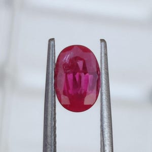 Puede incluir: Una gema ovalada de color rojo vibrante sostenida en unas pinzas. La gema tiene un tono rojo intenso y una apariencia ligeramente translúcida.
