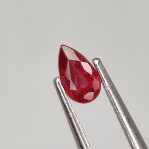 Op de afbeelding: Een peervormige rode edelsteen vastgehouden door een pincet. De edelsteen is gefacetteerd en heeft een dieprode kleur.