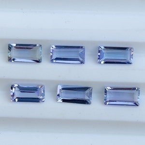 Könnte beinhalten: Sechs rechteckige Edelsteine im Baguetteschliff mit einem durchscheinenden, hellen azurblauen Farbton. Jeder Stein hat facettierte Kanten, die das Licht reflektieren, und ist in zwei horizontalen Reihen zu je drei auf einer weißen, gerillten Oberfläche angeordnet.
