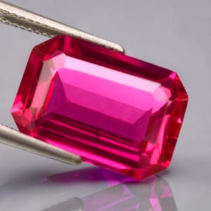 May include: A vivid, rectangular-cut, fuchsia gemstone is held by silver tweezers. The gem's facets reflect light, highlighting its clarity and rich colour. The gemstone is a deep pink hue, possibly a ruby or similar precious stone.