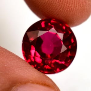 May include: A faceted, round-cut ruby gemstone. The gem is a deep, vibrant red colour with multiple facets that reflect light. The ruby is held between two fingers against a white background.