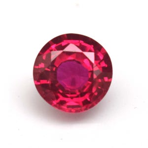 May include: A round, faceted ruby gemstone. The gem is a deep, vibrant red color with a lighter, almost pink center. The facets create a sparkling effect, and the gem is set against a white background.