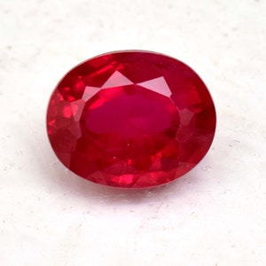 Könnte beinhalten: Ein facettierter, oval geformter Rubin-Edelstein. Der Edelstein hat eine tiefe, satte rote Farbe mit einer brillanten, reflektierenden Oberfläche. Der Stein liegt vor einem weißen Hintergrund.