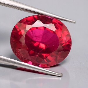 Peut inclure: Une pierre précieuse rouge vif de taille ovale tenue par une pince en métal argenté. La pierre présente une surface à facettes qui reflète la lumière, mettant en valeur une teinte cramoisie profonde avec une clarté interne et un aspect poli.