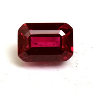 May include: A faceted, rectangular ruby gemstone with a deep red hue. The gem has a classic emerald cut with beveled corners, reflecting light and showcasing its clarity. The ruby is set against a white background.
