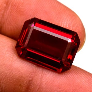 Puede incluir: Una gema roja oscura tallada en forma rectangular y facetada. La gema tiene una superficie pulida y bordes afilados. El color es un rojo rico y saturado, con reflejos internos visibles. La gema es sostenida por los dedos de una persona.