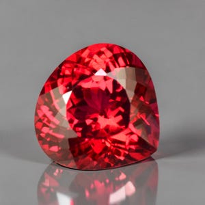 Könnte beinhalten: Ein facettierter, herzförmiger Edelstein in leuchtendem Rot. Die Oberfläche des Edelsteins reflektiert das Licht und zeigt seine komplizierten Facetten und seine innere Brillanz. Der Edelstein ist vor einem neutralgrauen Hintergrund platziert.