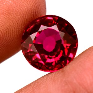 May include: A faceted, round, ruby gemstone. The gem is a deep, saturated red color with multiple facets that reflect light. The gemstone is held between two fingers against a white background.