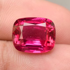 Op de afbeelding: Een levendige, kussengeslepen edelsteen met een diepe, verzadigde magenta-rode kleur. De steen heeft een gefacetteerd oppervlak dat licht reflecteert, wat zorgt voor een briljante, sprankelende uitstraling met scherpe geometrische randen.