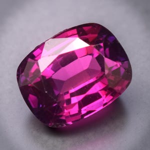 May include: A faceted, cushion-cut gemstone with a vibrant magenta hue. The gem's facets reflect light, creating a sparkling effect. The stone's shape is rectangular with rounded corners, and it appears to be set against a neutral background.