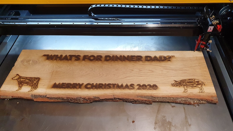 Peut inclure: Une pancarte en bois avec le texte "What's for dinner Dad?" et "Merry Christmas 2020" br&ucirc;l&eacute; &agrave; la surface. La pancarte pr&eacute;sente &eacute;galement une vache et un cochon avec des lignes de boucherie dessin&eacute;es dessus.
