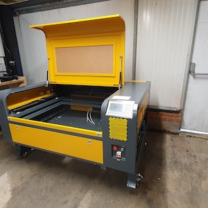 Peut inclure: Une d&eacute;coupeuse laser jaune et grise avec le couvercle ouvert, r&eacute;v&eacute;lant le lit de coupe. La machine est sur roulettes et poss&egrave;de un panneau de commande sur le c&ocirc;t&eacute;. Le texte "Push bar to open" est visible sur une porte en arri&egrave;re-plan.