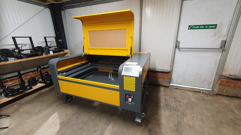 Peut inclure: Une d&eacute;coupeuse laser jaune et grise avec le couvercle ouvert, r&eacute;v&eacute;lant le lit de coupe. La machine est sur roulettes et poss&egrave;de un panneau de commande sur le c&ocirc;t&eacute;. Le texte "Push bar to open" est visible sur une porte en arri&egrave;re-plan.