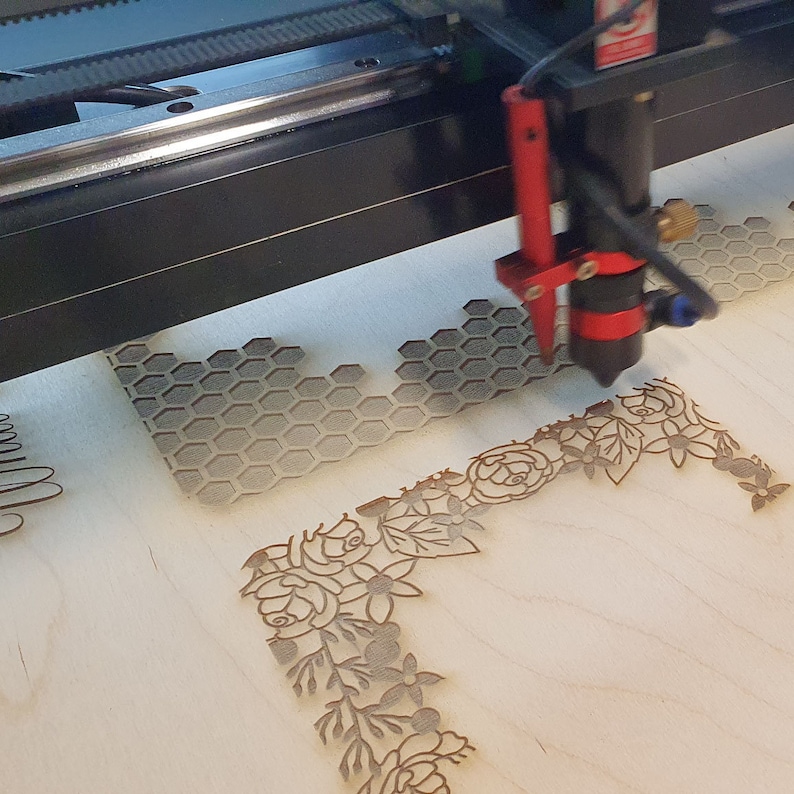 Peut inclure: Une d&eacute;coupeuse laser grave un motif sur un morceau de bois. Le motif pr&eacute;sente un motif en nid d'abeille et une bordure florale.