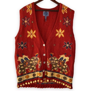 May include: Red knitted vest with a V-neck and button closure. The vest features an embroidered design of elephants and flowers. The vest has a beaded trim around the bottom.