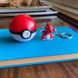 Può includere: Un giocattolo Poké Ball rosso e bianco accanto a un portachiavi Charmander rosso. La Poké Ball è rotonda con una fascia nera e un cerchio bianco. La figura di Charmander è rossa con la pancia bianca e attaccata a un portachiavi argentato.
