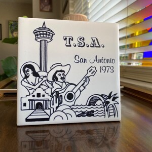 May include: A white ceramic tile with a black and white illustration of a cityscape, a person playing a guitar, and the text "T.S.A. San Antonio 1973".