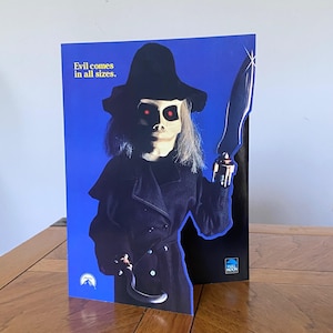 May include: A blue and black cardboard stand-up of a puppet with a black hat, a black coat, and a knife. The puppet has red eyes and a creepy smile. The text "Evil comes in all sizes." is printed on the stand-up.