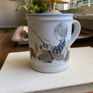 May include: White ceramic mug with a blue rim. The mug features a hand-painted illustration of a boy fishing with a dog by his side. The boy is wearing a blue and white striped shirt and brown pants. The scene is set in a field with green grass and yellow flowers.
