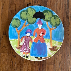 May include: A hand-painted ceramic plate featuring a whimsical scene of two figures under a black umbrella, with trees in the background. The figures are dressed in colorful clothing, with a blue sky and green grass. The plate has a black string for hanging.