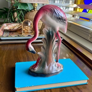 May include: A ceramic flamingo figurine with a pink body, neck, and head, and a black beak. The flamingo is perched on a gray and pink base with green accents. The figurine is placed on a blue book.
