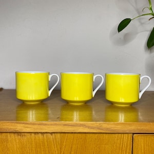 Puede incluir: Tres tazas de cerámica amarilla con bordes blancos. Las tazas están sobre una superficie de madera.