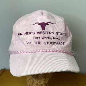 Könnte beinhalten: Rosa Cord-Baseballkappe mit einem lila bestickten Logo, das "Fincher's Western Stores Fort Worth, Texas "At The Stockyards" lautet.
