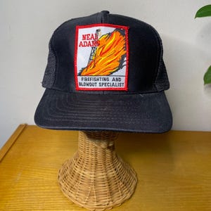 May include: Black trucker hat with a red, white, and yellow patch that reads "Neal Adams Firefighting and Blowout Specialist".