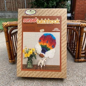 May include: A Sunset Latchhook kit featuring a colorful hot air balloon design, with the text "FREEDOM'S FLIGHT". The kit includes a finished size of 12" x 14". Also shown are yellow flowers in a glass vase and a white figurine.