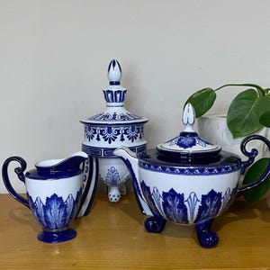 May include: A white and blue porcelain tea set featuring a teapot, creamer, and lidded sugar bowl. The set has a detailed blue leaf pattern and is elevated on small feet. The teapot has a curved handle and spout.