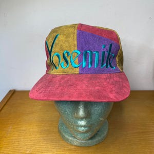 Peut inclure: Casquette de baseball colorée avec le mot "Yosemite" brodé en turquoise. La casquette a une visière rouge et un motif patchwork en or, violet et rouge. Le chapeau est présenté sur une tête de mannequin.